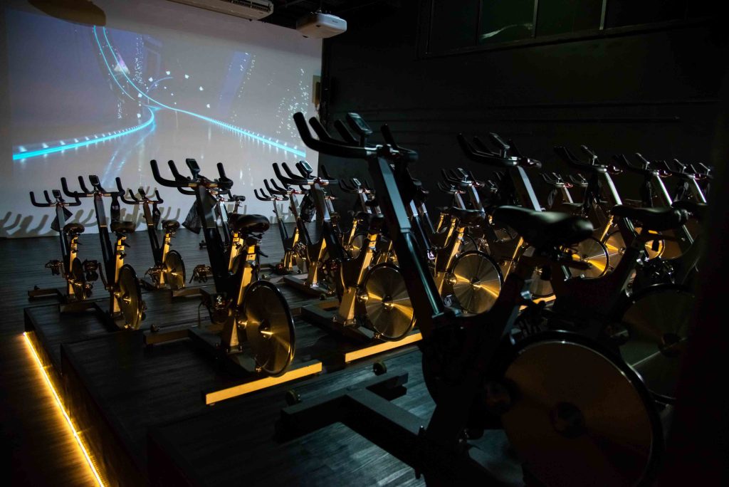 Sala de spinning da Cia Athletica SJC com bicicletas ergométricas organizadas em fileiras, iluminação baixa e projeção audiovisual em uma grande tela ao fundo, criando um ambiente moderno e imersivo para treinos indoor.