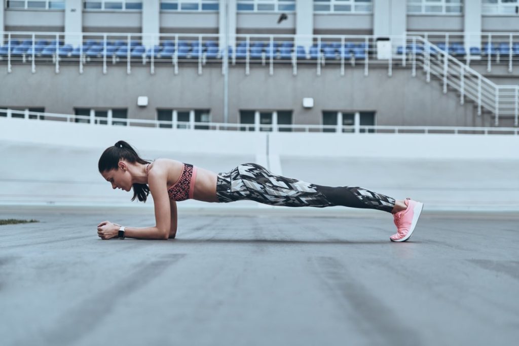 uma mulher está realizando o exercício de prancha, mantendo o corpo em linha reta, apoiada nos antebraços e nas pontas dos pés