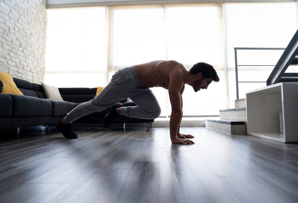 homem sem camisa realizando um exercício de "mountain climbers" em casa, um movimento que trabalha intensamente a região abdominal e os músculos centrais do corpo.