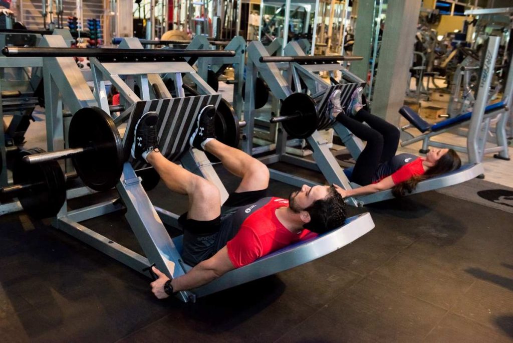 vemos duas pessoas realizando treino de pernas na academia utilizando o aparelho leg press