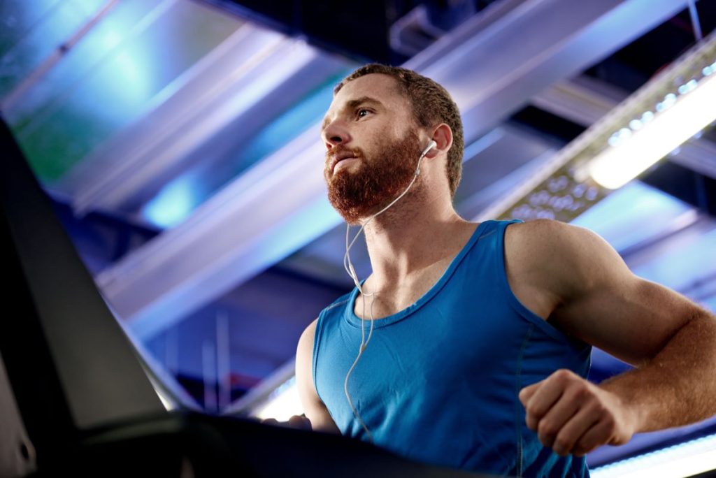 homem correndo em uma esteira em uma academia, usando regata azul e fones de ouvido.