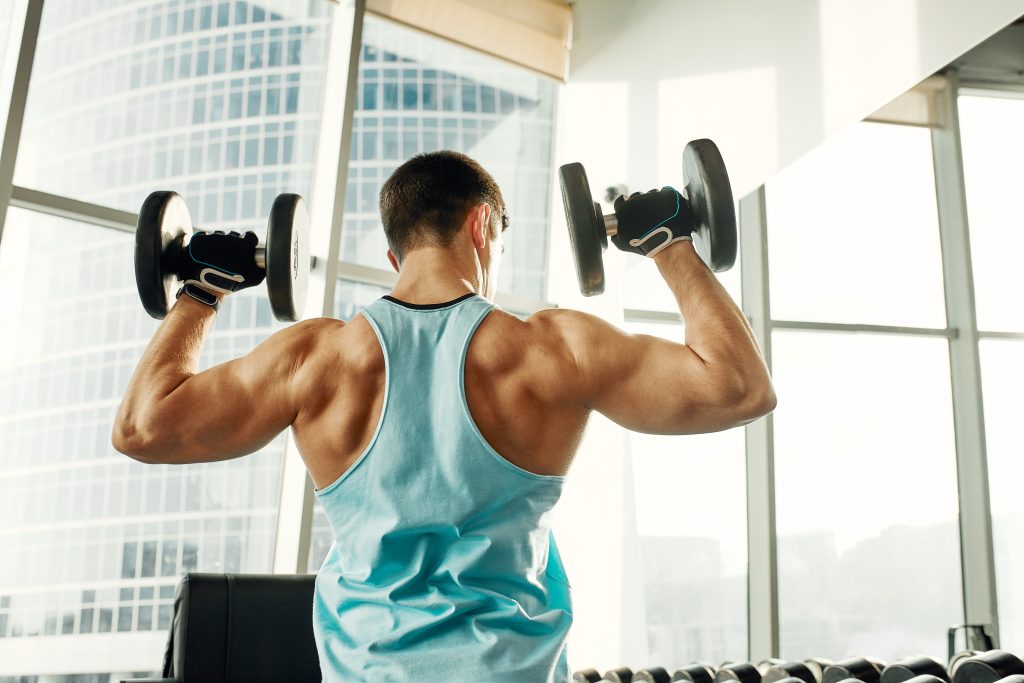 Pessoa realizando desenvolvimento de ombros com halteres em academia, fortalecendo deltoides e membros superiores durante treino de musculação.