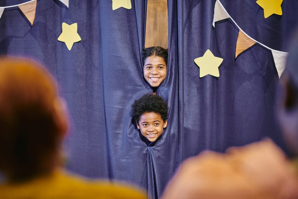 Crianças sorridentes participando de apresentação de circo com cortina azul e estrelas amarelas, desenvolvendo coordenação motora e criatividade na escola de circo infantil.