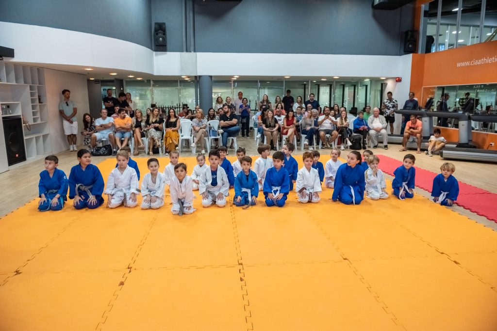 Alunos de judô da Cia Athletica SJC sentados em fileiras sobre o tatame durante apresentação infantil, com pais e responsáveis assistindo ao fundo em ambiente de academia.