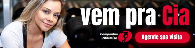 Mulher sorridente apoiada em equipamentos de musculação na Cia Athletica São José dos Campos, com halteres ao fundo e o convite "Vem pra Cia – Agende sua visita" em destaque.