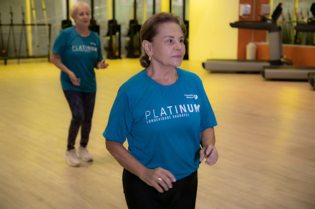 Duas mulheres idosas participam de uma atividade física em uma academia da Cia Athletica São José dos Campos, caminhando em um espaço amplo e iluminado enquanto usam camisetas azuis do programa Platinum, voltado à longevidade saudável.