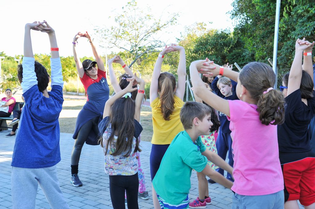 Instrutora conduz atividade de alongamento com grupo de crianças ao ar livre durante o Programa Kids SJC da Cia Athletica SJC, incentivando desenvolvimento motor, coordenação e prática de atividade física infantil.
