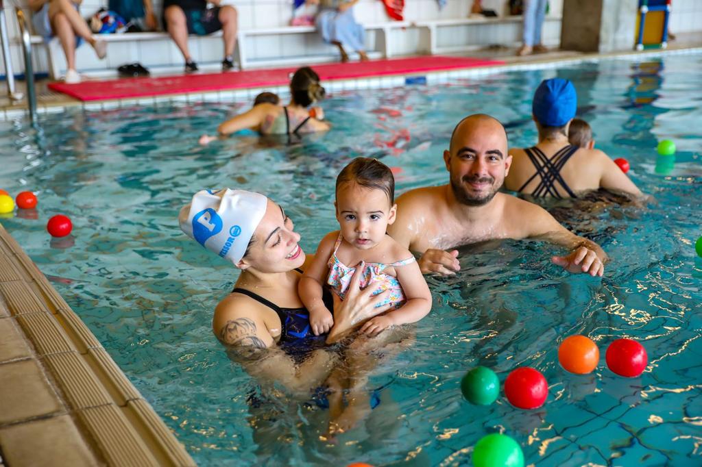 Mulher segura um bebê dentro de uma piscina coberta enquanto um homem permanece ao lado, em atividade aquática com outras famílias e bolas coloridas ao redor.