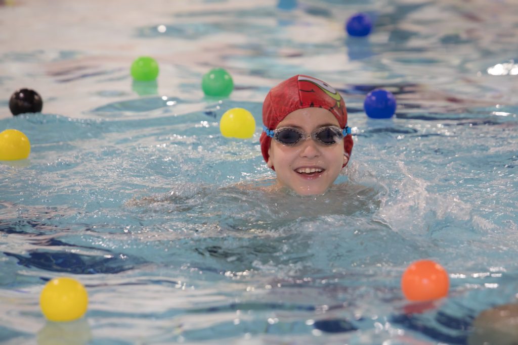 Criança nada em piscina usando touca vermelha e óculos de natação, cercada por bolas coloridas durante uma atividade aquática infantil.