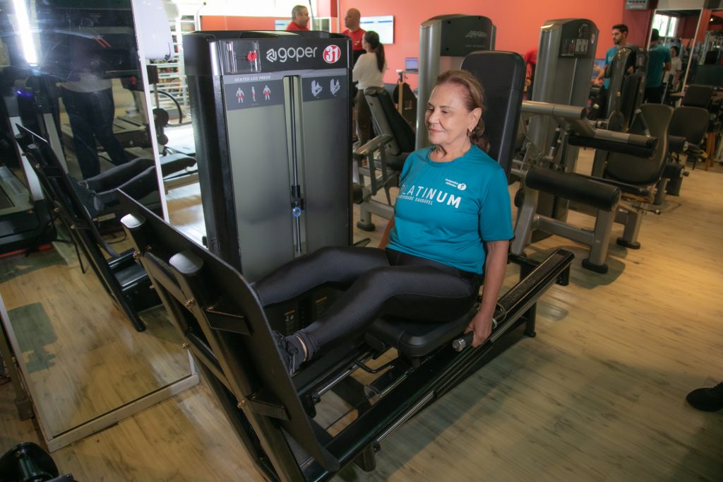 Mulher idosa realiza exercício de leg press em aparelho de musculação dentro de uma academia, usando camiseta azul e calça preta.